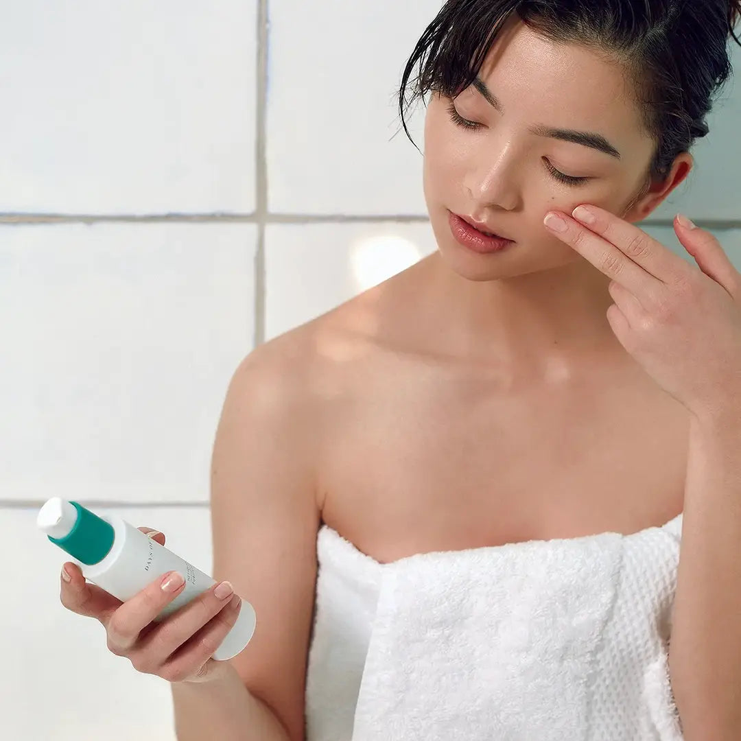 fille peau blanche coréenne qui tient le nettoyant purifiant visage dans une salle de bain à carreaux avec serviette autour du corps