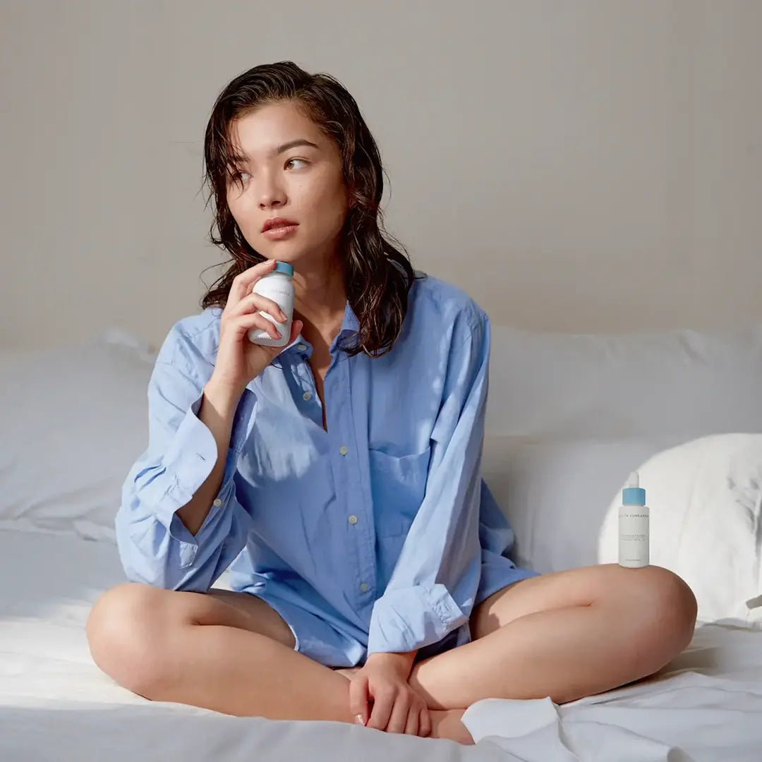 fille eurasienne assise sur un lit vêtue d’une chemise bleu qui tient dans sa main le complément alimentaire sérénité days of confidence et sur son genoux l’huile apaisante visage